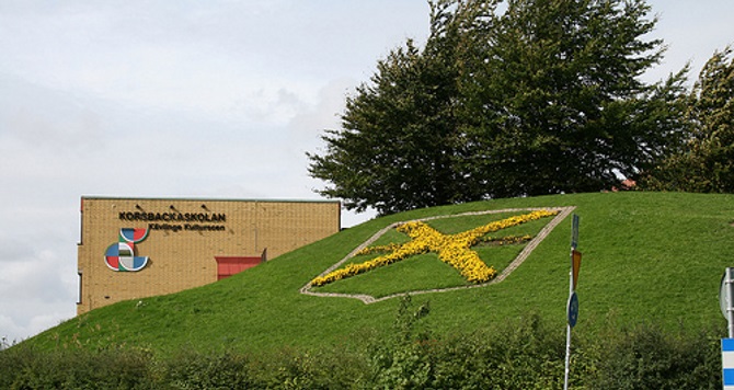 Korsbackaskolans gula tegelgavel och kommunens vapen på en grönskande kulle