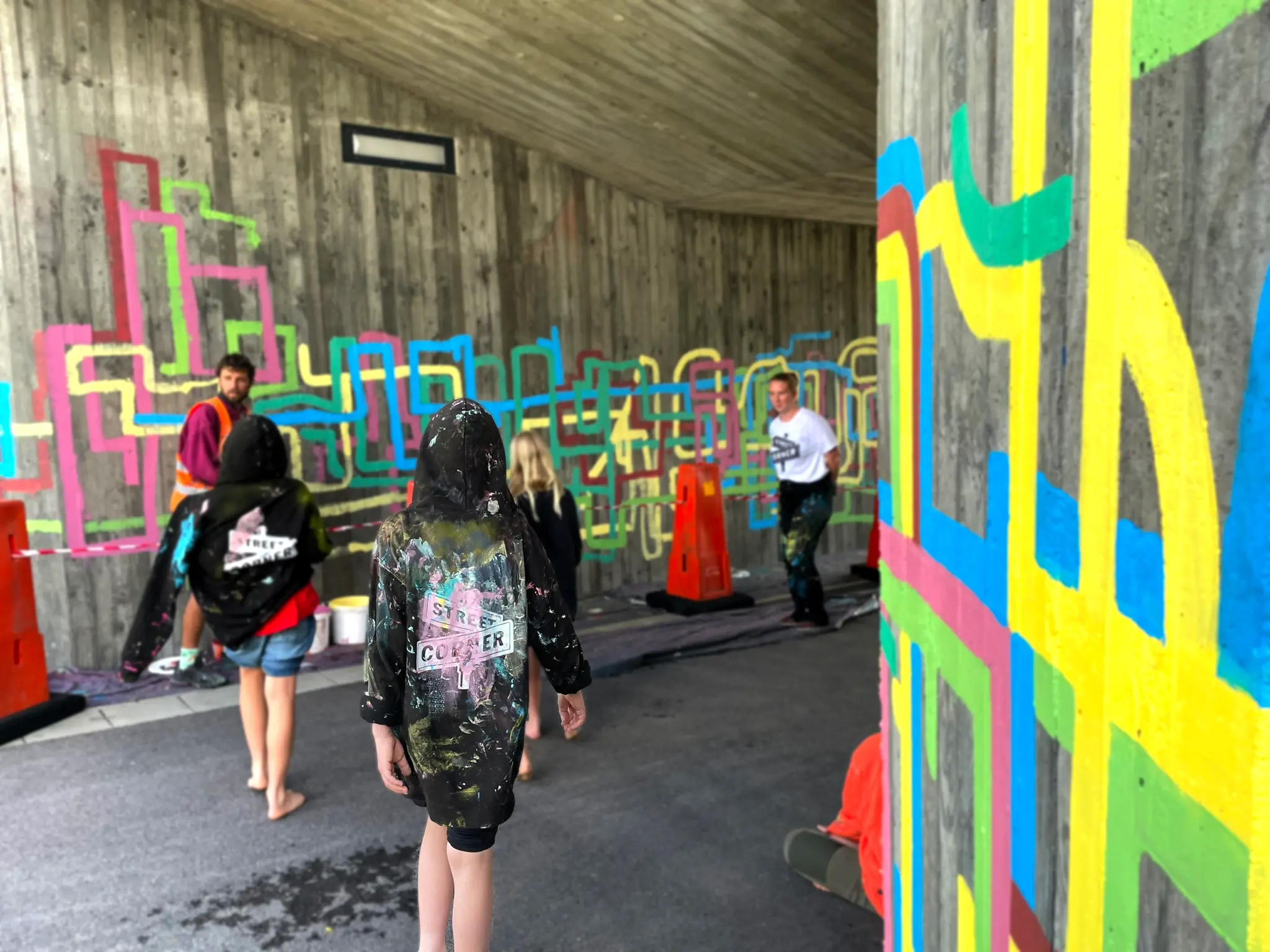 Ryggtavla på ett barn med svart huvtröja med färgrester på i en gångtunnel med färgglad konst på väggarna.