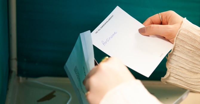 En hand stoppar i en valsedel i ett valkuvert. 