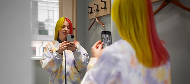 Ung, vuxen kvinna med gult och rosa hår provar en jacka och tar en selfie i spegeln i en provhytt.